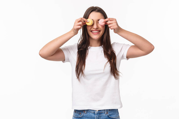 Waist-up portrait of funny and cute, adorable young woman holding two desserts macarons over eyes and smiling, playing with food, fool around, standing upbeat over white background - Foto, Bild