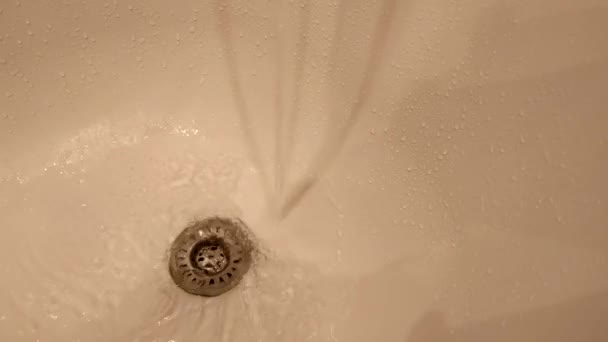 A csapból víz folyik a fürdőkádba és a lefolyóba. Vízcseppek a fürdő belsejében. Top kilátás a fürdőkádra. - Felvétel, videó