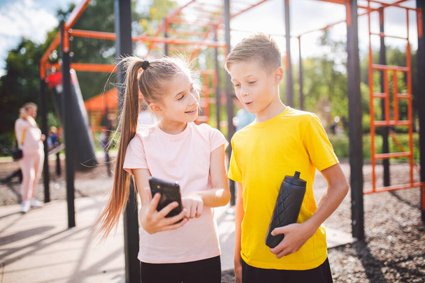 Technik und Sport für Kinder. Zwei Teenager mit Smartphone und Online-Workout-App. Zwillinge Jungen und Mädchen sehen während der Pause vom Sportunterricht in der Schule in der Sporthalle ein Handy-Video. - Foto, Bild
