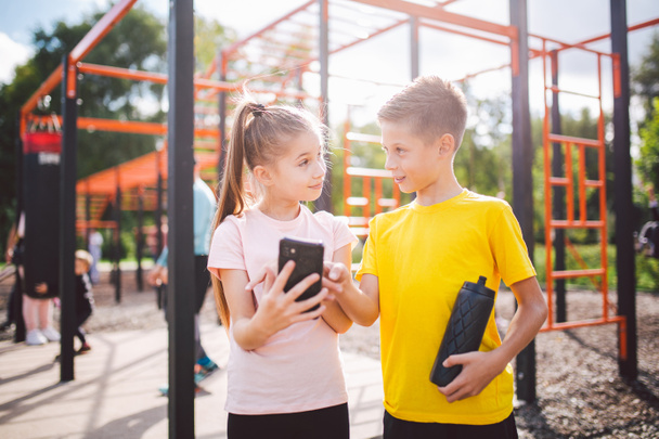 Καυκάσια παιδιά έφηβοι χρησιμοποιούν το κινητό τηλέφωνο στην προπόνηση στο γυμναστήριο του δρόμου. Διαφορετικές δίδυμες παρακολουθούν μαθήματα βίντεο με ασκήσεις γυμναστικής. Αδελφός και αδελφή χρησιμοποιούν την εφαρμογή άσκησης στο smartphone από μόνοι τους. - Φωτογραφία, εικόνα