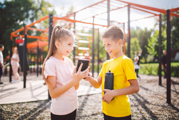 白人の子供たちはストリートジムのワークアウトで携帯電話を使用します。体操でビデオレッスンを見て多様な双子。兄と妹は自分でスマートフォンで運動アプリを使用しています. - 写真・画像
