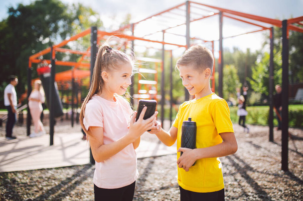 Technology and sport for children. Two teenage kids using smartphone and online workout app. Twins boy and girl watching video on phone during break from sports lesson at school at outside gym. - Foto, afbeelding