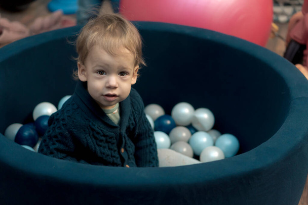 Childhood, infants, development concept - portrait of small preschool minor, fair-haired child boy of 1-2 years old in arena with small colored balls. Kid without emotions sad or scared in playroom - Foto, imagen