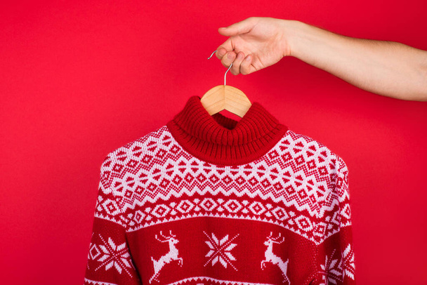 Feo suéter de Navidad concepto de fiesta. Foto de cerca de hombres sosteniendo percha con suéter de invierno rojo con ciervos aislados sobre fondo rojo - Foto, Imagen