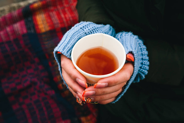 Μια Ευρωπαία που κρατάει ένα φλιτζάνι τσάι σε κρύα χέρια σε καρό φόντο. Θέρμανση χέρια με ζεστό τσάι και πουλόβερ μανίκια. - Φωτογραφία, εικόνα