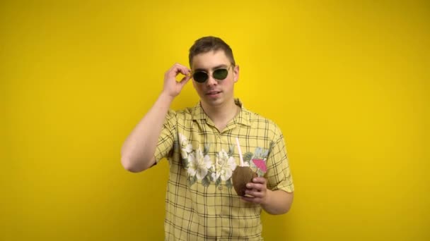 Un hombre con gafas de sol y una camisa hawaiana se quita las gafas y tiene una piña colada de coco en la mano. Tiro en el estudio sobre un fondo amarillo. - Imágenes, Vídeo