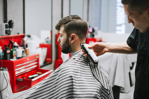 Männliche Friseur schafft eine stilvolle Frisur für einen erwachsenen Mann mit Bart in einem leichten Friseurladen. Friseur schneidet den Kunden mit einem Haarschneider. Barboshop-Konzept. - Foto, Bild
