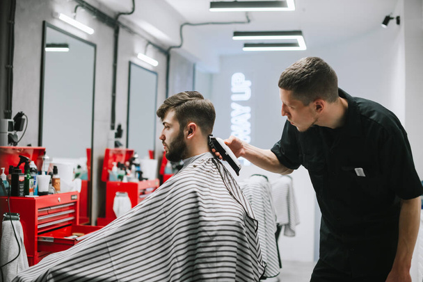 Professional barber with a hair clipper in a back hair cuts an adult client's hair with a beard. Focused male hairstylist creates a stylish hairstyle for a man in a light hairdresser. - Photo, Image