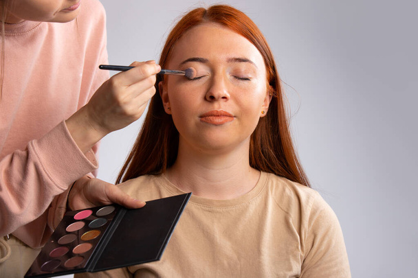 A make-up artist holds a palette in his hands and does make-up to a red-haired girl. High quality photo - Foto, Imagen