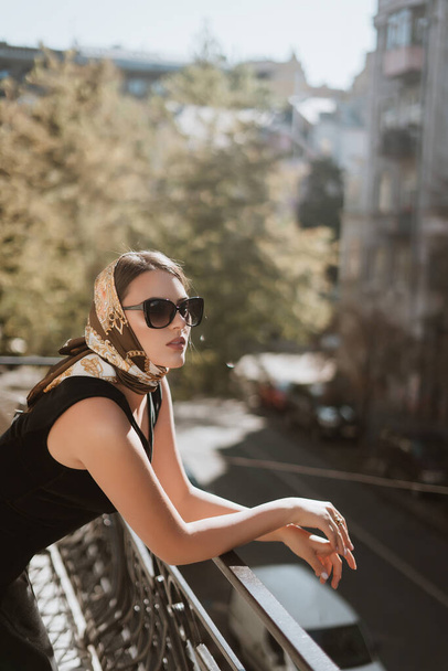 Outdoor close up fashion portrait of elegant woman wearing trendy colorful head scarf, black dress, black eye sunglasses. lady resting on the balcony. vintage style - Fotografie, Obrázek