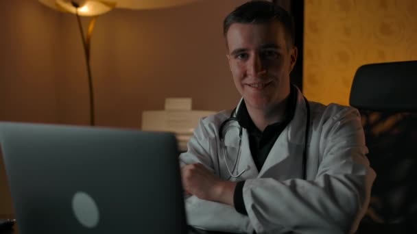 Uśmiechnięty lekarz w szlafroku za laptopem uśmiechnięty do kamery - Materiał filmowy, wideo