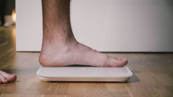 Человек наступает на цифровые стеклянные весы, чтобы проверить вес. Мужчина взвешивает на полу весы в бытовой комнате, закрывает вид на ноги. Мужские ноги на электронных весах. Спортивная концепция. Похудение и здоровый образ жизни. - Фото, изображение