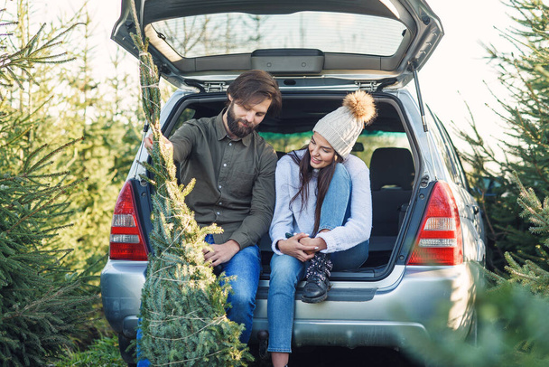 Couple in love loading freshly cut down christmas tree into a trunk of their car. Young family preparing to holidays. - Fotoğraf, Görsel