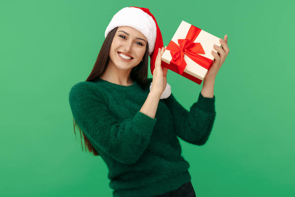 Gleeful mladá brunetka žena nosit Santa Claus klobouk a zelený svetr držení dárkové krabice přes zelené pozadí. - Fotografie, Obrázek