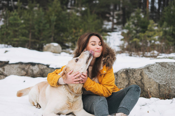 Joven mujer sonriente en chaqueta amarilla con perro blanco de gran clase Labrador caminando en el bosque de invierno - Foto, Imagen
