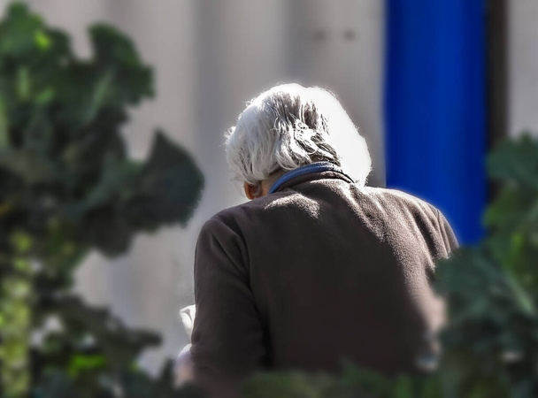 Eine alte Frau mit weißem Haar und brauner Bluse im Garten neben dem Haus - Foto, Bild