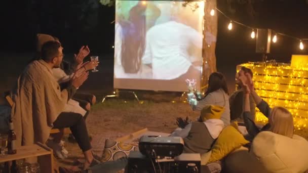 Друзья смотрят фильм на открытом воздухе через киноэкран и проектор в летний вечер аплодируют актерам в замедленной съемке - Кадры, видео