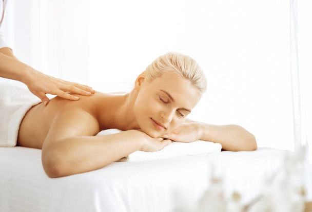 Beautiful blonde woman enjoying back massage with closed eyes in sunny spa salon. Beauty concept - 写真・画像