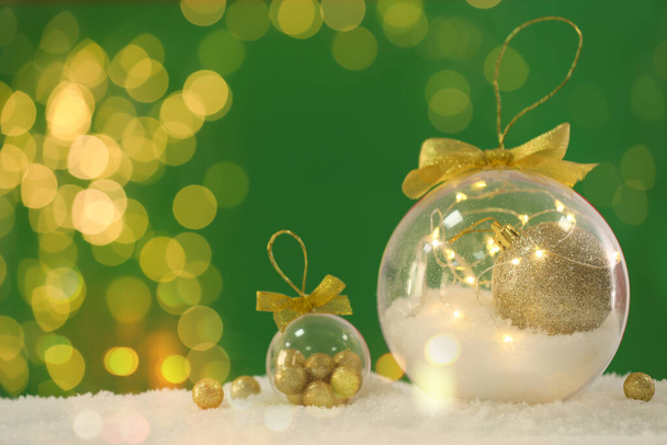 Beautiful transparent Christmas ornaments with golden bows on snow against green background, bokeh effect. Space for text - Foto, afbeelding