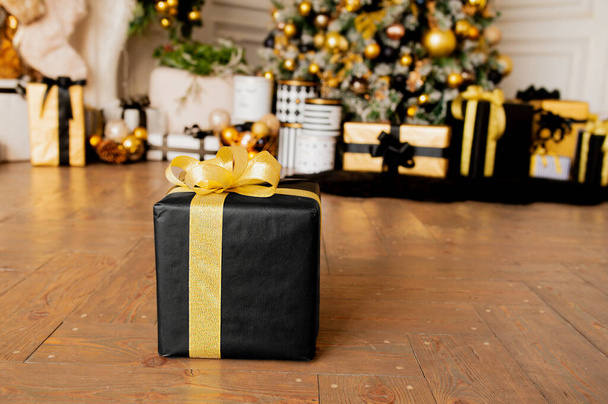 μαύρο κουτί δώρου με χρυσή κορδέλα σε ξύλινο πάτωμα στο δωμάτιο με χριστουγεννιάτικη διακόσμηση σε χρυσό και μαύρο χρώμα: Χριστουγεννιάτικο δέντρο με μπάλες και γιρλάντες και πολλά δώρα πακέτο κάτω από αυτό, επιλεκτική εστίαση - Φωτογραφία, εικόνα