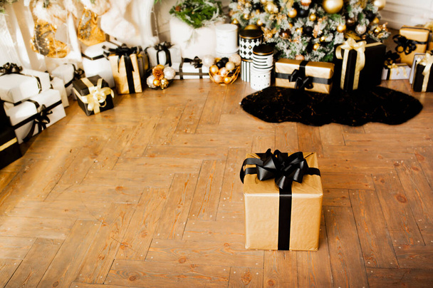 χρυσό κουτί δώρου με μαύρη κορδέλα σε ξύλινο πάτωμα στο δωμάτιο με χριστουγεννιάτικη διακόσμηση σε χρυσό και μαύρο χρώμα: Χριστουγεννιάτικο δέντρο με μπάλες και γιρλάντες και πολλά δώρα συσκευασμένο κάτω από αυτό, επιλεκτική εστίαση - Φωτογραφία, εικόνα