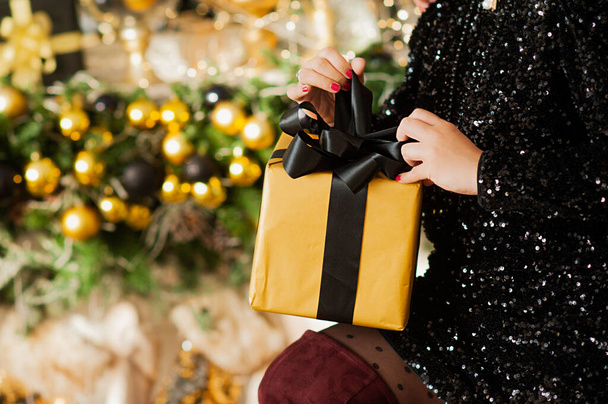 Hände der Frau in schwarzem Paillettenkleid mit großem goldenem Geschenk mit schwarzer Schleife, Girlanden und Bokeh auf weihnachtlichem Interieur auf Hintergrund, selektiver Fokus - Foto, Bild
