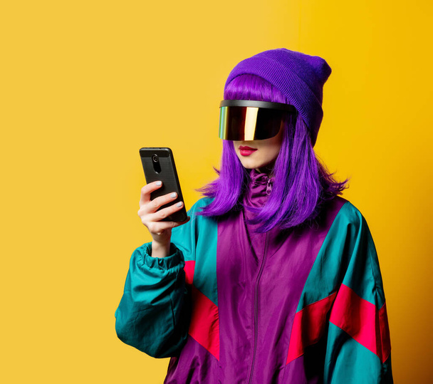 VR gözlüklü ve sarı arka planda cep telefonu olan 80 'lerin eşofmanlı stil kadını. - Fotoğraf, Görsel