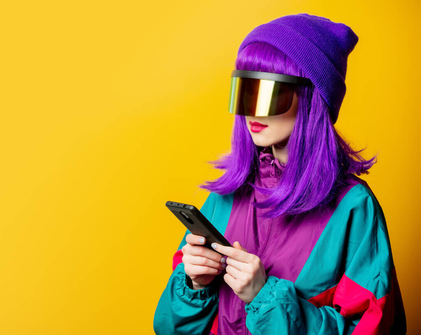Stijl vrouw in VR bril en 80s trainingspak met mobiele telefoon op gele achtergrond - Foto, afbeelding