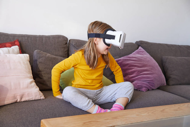 six years old child sitting on a brown sofa at home watching virtual reality headset glasses and talking - Photo, Image