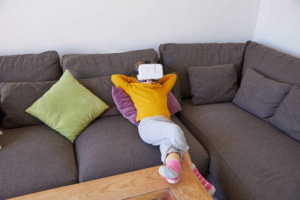enfant de six ans couché confortablement sur un canapé brun avec lunettes de casque de réalité virtuelle - Photo, image