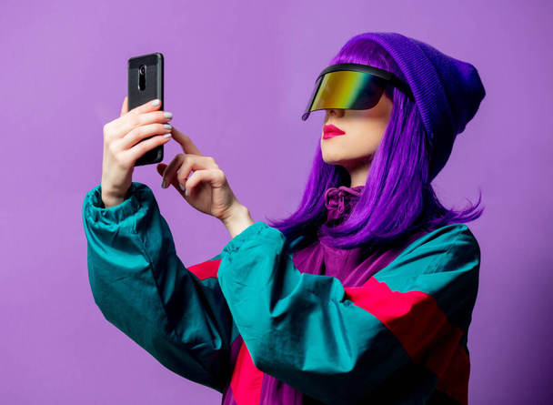 紫の背景に携帯電話でVRメガネと80年代のトラックスーツのスタイルの女性 - 写真・画像