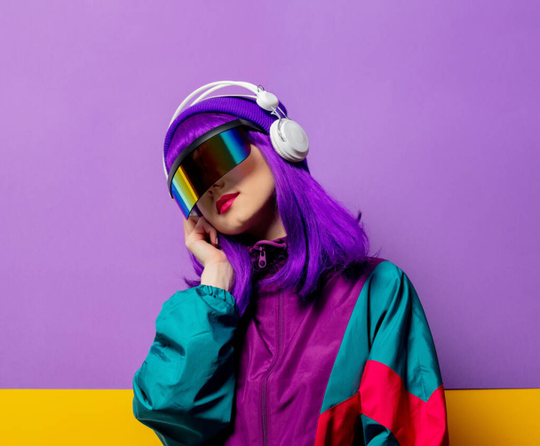 Stijl vrouw in VR bril en 80s trainingspak met hoofdtelefoon op violette achtergrond - Foto, afbeelding