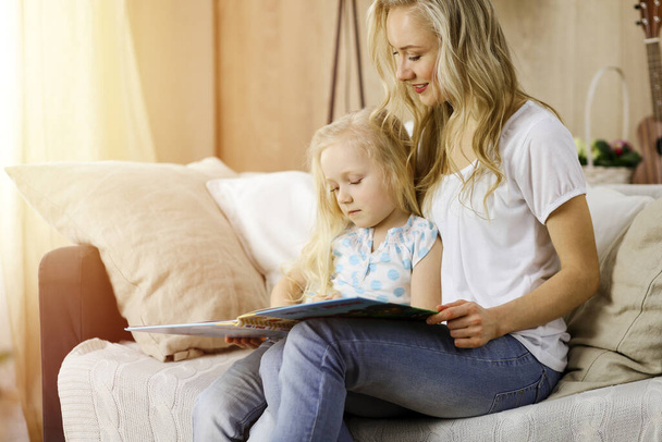 Happy family. Blonde young mother reading a book to her cute daughter while sitting at wooden floor in sunny room. Motherhood concept - Foto, imagen