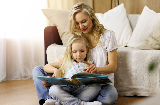 Happy family. Blonde young mother reading a book to her cute daughter while sitting at wooden floor in sunny room. Motherhood concept - 写真・画像
