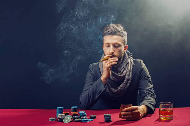 Stylish bearded Man in suit and scarf playing in dark casino, smoking cigar, drink whiskey - Foto, Bild