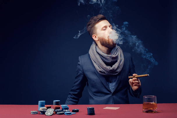 Stylish bearded Man in suit and scarf playing in dark casino, smoking cigar, drink whiskey - Foto, imagen