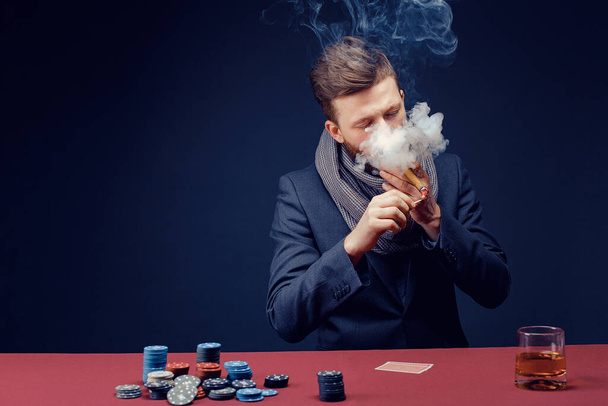 Stylish bearded Man in suit and scarf playing in dark casino, smoking cigar, drink whiskey - Foto, Bild