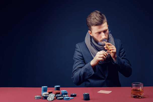 Stylish bearded Man in suit and scarf playing in dark casino, smoking cigar, drink whiskey - 写真・画像