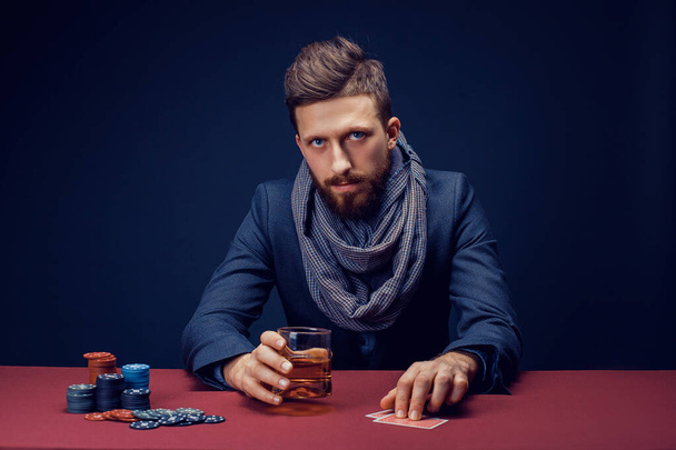 Stylish bearded Man in suit and scarf playing in dark casino, smoking cigar, drink whiskey - Foto, afbeelding