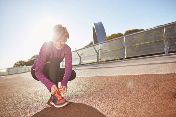 Готовься. Полнометражный снимок активной взрослой женщины в спортивной одежде, завязывающей шнурки на спортивных кроссовках, готовящейся к выходу на улицу в солнечный день - Фото, изображение
