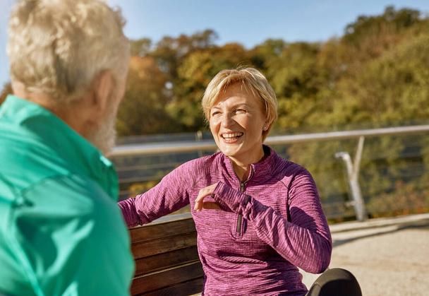 Glückliches, sportliches Seniorenpaar, das nach dem Training im Freien auf der Bank sitzt. Ältere Frau lächelt Mann an, während sie etwas diskutiert - Foto, Bild
