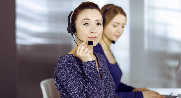 Two busineswomen have conversations with the clients by headsets, while sitting at the desk in a modern office. Diverse people group in a call center. Telemarketing and customer service - Fotografie, Obrázek