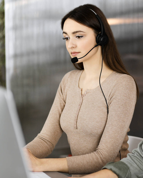 Two young people in headsets are talking to the clients, while sitting at the desk in a modern office. Focus on woman. Call center operators at work - Foto, Imagen