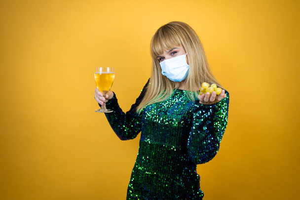 Beautiful blonde woman wearing a fancy dress and medical mask over yellow background holding a glass of champagne and grapes. New Years Eve celebration - Fotografie, Obrázek