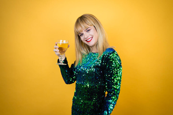 Sarı arka planda süslü bir elbise giyen güzel sarışın kadın elinde bir bardak şampanya tutuyor. Yeni Yıl kutlamaları - Fotoğraf, Görsel