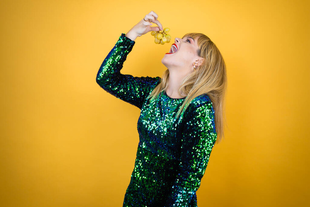 Beautiful blonde woman wearing a fancy dress over yellow background holding a bunch of grapes. New Years Eve celebration - Foto, afbeelding