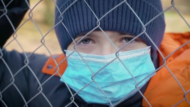 Trauriger Junge in medizinischer Schutzmaske zur Coronavirus-Prävention, der während der Quarantäne der Epidemie hinter Netzen steht - Filmmaterial, Video