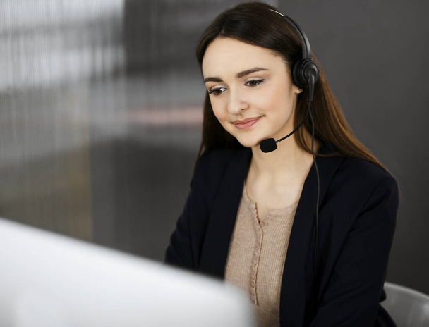 Young friendly girl in headsets is talking to a firms client, while sitting at the desk in a modern office together with her colleague. Call center operators at work - Fotó, kép