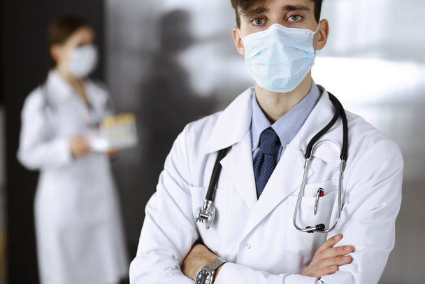 Fiatal férfi orvos áll keresztbe tett karokkal a klinikán. Okos orvos orvosi védőmaszkot visel a modern kórházban. Gyógyszer és a Corona vírus fogalma - Fotó, kép