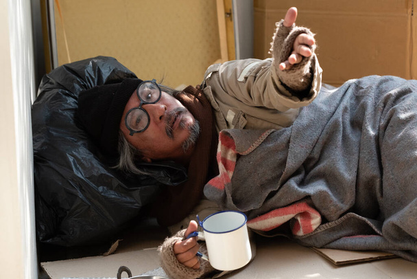 Ένας άστεγος δέχεται μια μάσκα προσώπου από έναν περαστικό, καθώς είναι άνεργος και πεινασμένος. Μένει στο πεζοδρόμιο λόγω των 19 πανδημιών.. - Φωτογραφία, εικόνα